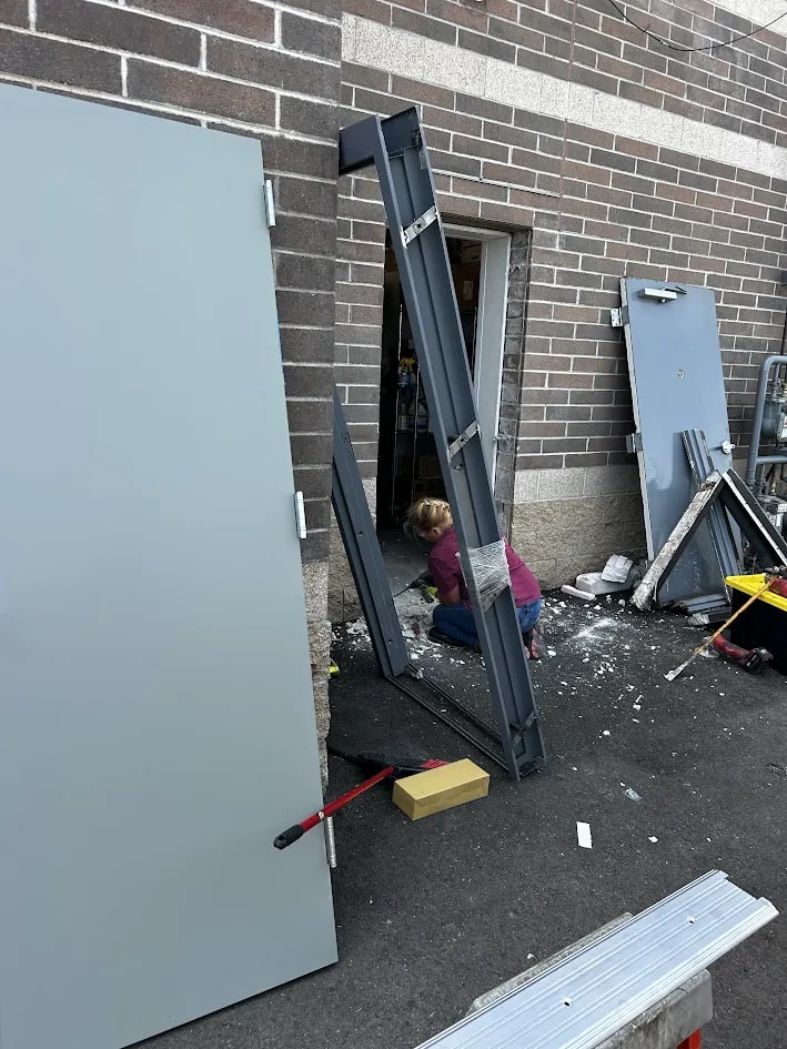 Two new grey hollow metal door leaves leaning against a brick wall beside a new knock-down steel frame, with a technician working at the opened wall