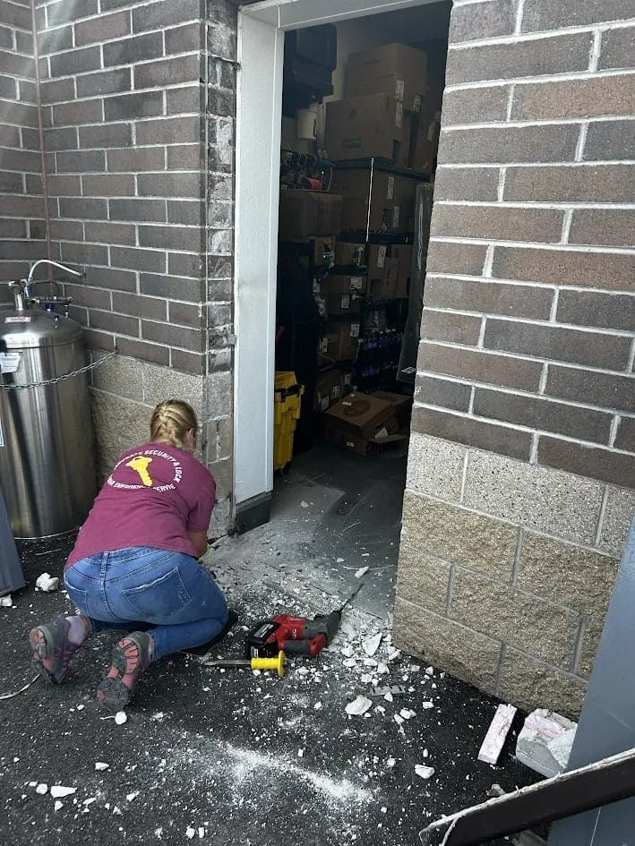 Technician kneeling at a commercial door opening with a power tool, debris on the ground and a storeroom visible behind