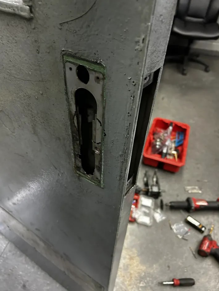 Steel door with a rectangular aperture cut at the lock case, exposing the internal mechanism, surrounded by tools and a red tray of fixings