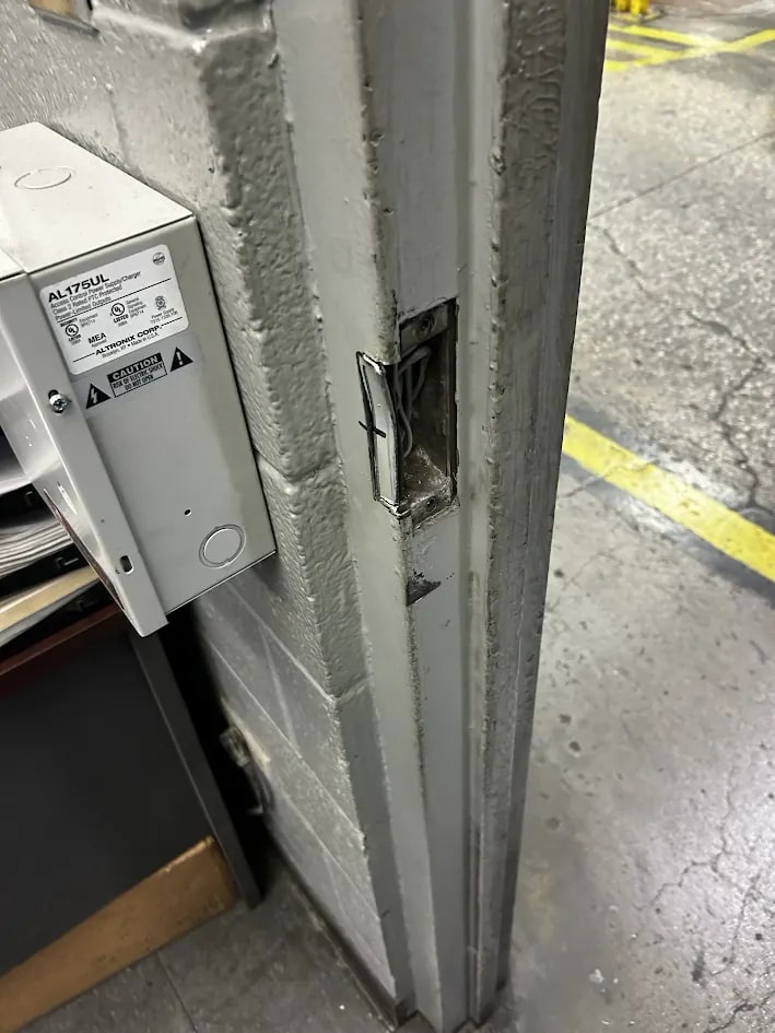 Steel door jamb cut open in a rectangle to accept an electric strike, with conductors visible in the cavity and a supply box on the wall to the left