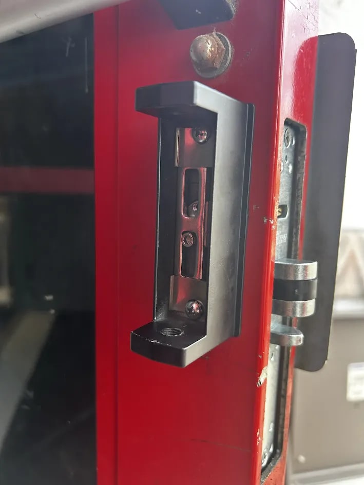 Heavy black strike plate screwed to a red painted steel frame, with the door edge and its latch plate alongside