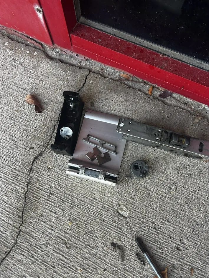 Lock hardware and fixings arranged on a concrete floor at the foot of a red door, including a lock body, black bracket and cylinder