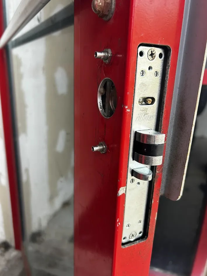 Edge of a red-framed glass door with the mortise lock faceplate off, threaded studs exposed and the cylinder hole open