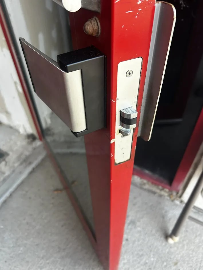 Black-backed paddle with a brushed face and a steel latch faceplate and cam at the edge of the red-framed glass door