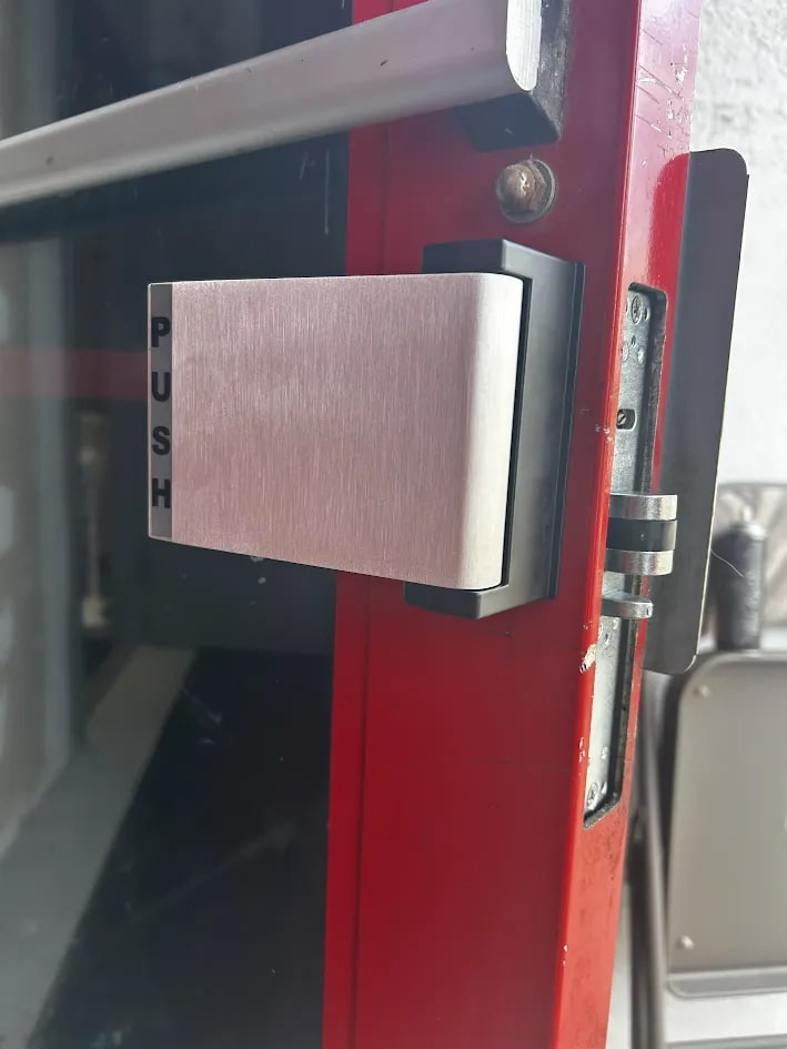 Large brushed stainless PUSH plate on a black mounting block, fixed to the stile of a red-framed glass door