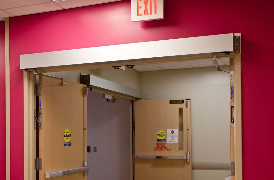 Wooden double doors with panic push bars and exit signs in a pink painted hallway