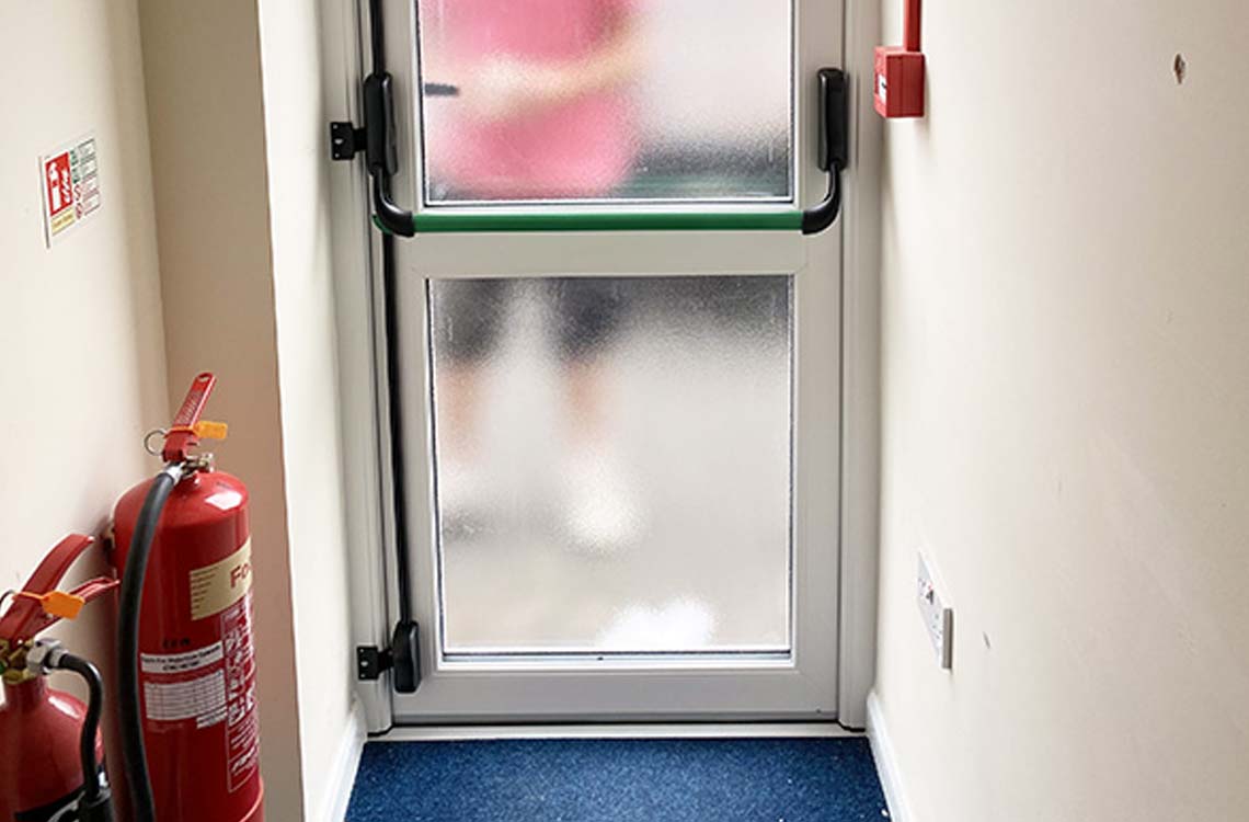 Metal framed frosted glass door with green push bar beside a red fire extinguisher in a corridor