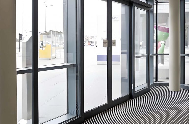 Black framed glass entrance doors with NHS sign, striped floor mat, yellow phone kiosk outside