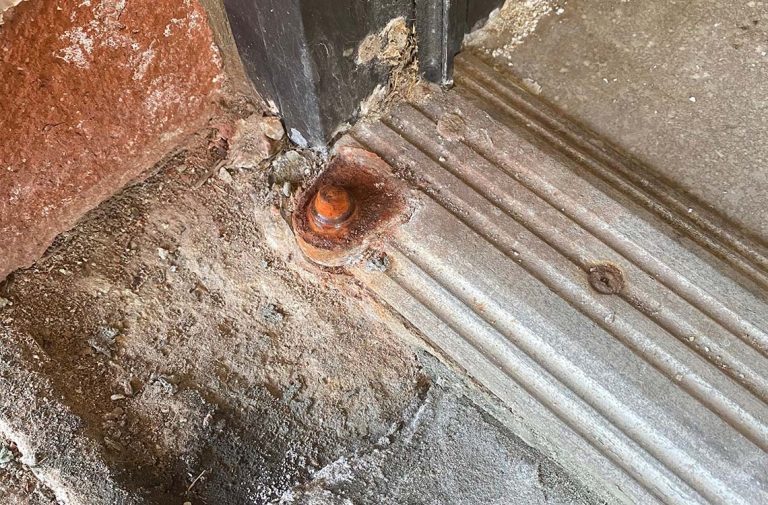 Rusted floor pivot hinge in threshold concrete beside black door frame