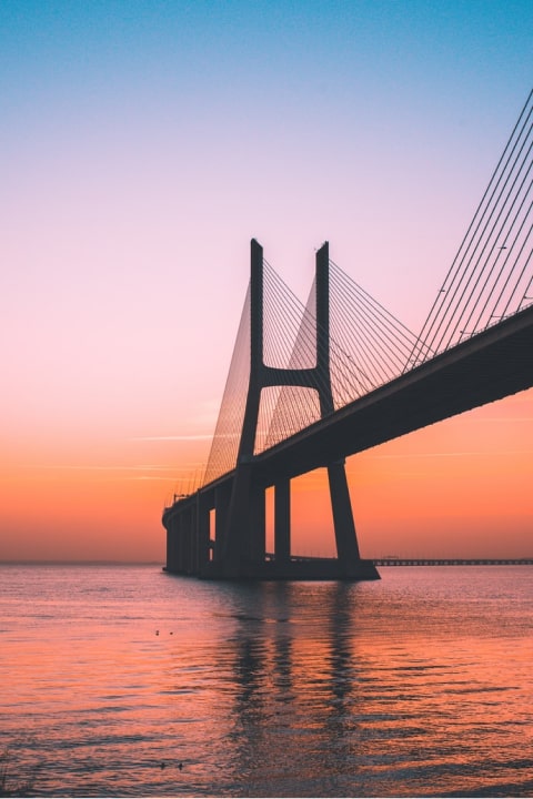 Cable-stayed bridge towers over calm water at orange sunset, city skyline beyond